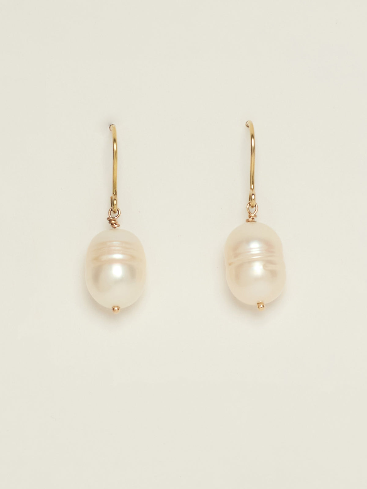 A pair of gold drop earrings featuring large, lustrous pearls with iridescent white and pink hues, displayed on a clean off-white background.