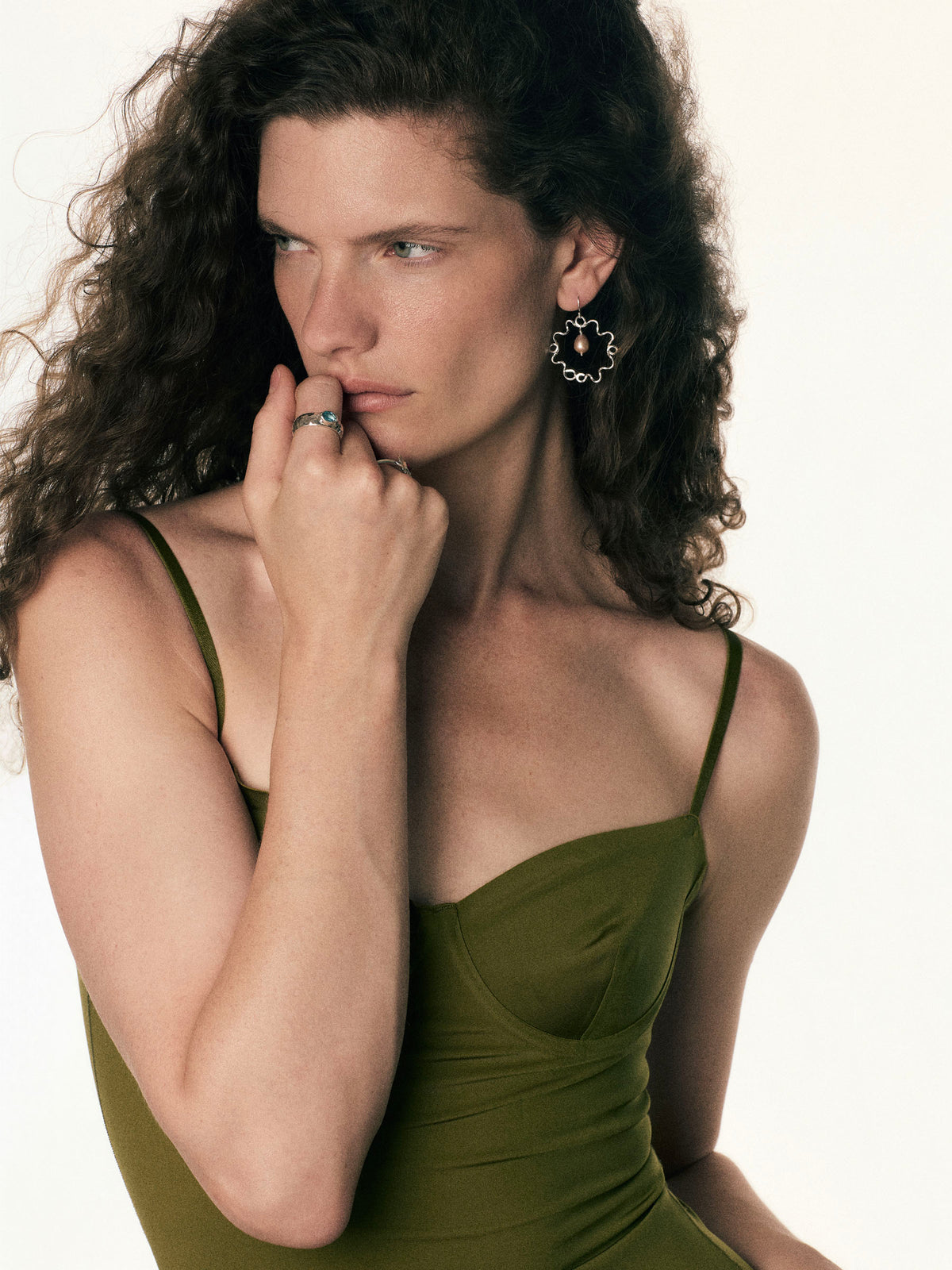A woman with long, curly dark brown hair wearing an olive green spaghetti-strap dress and a silver  hoop pearl earring, posing against a clean white background.
