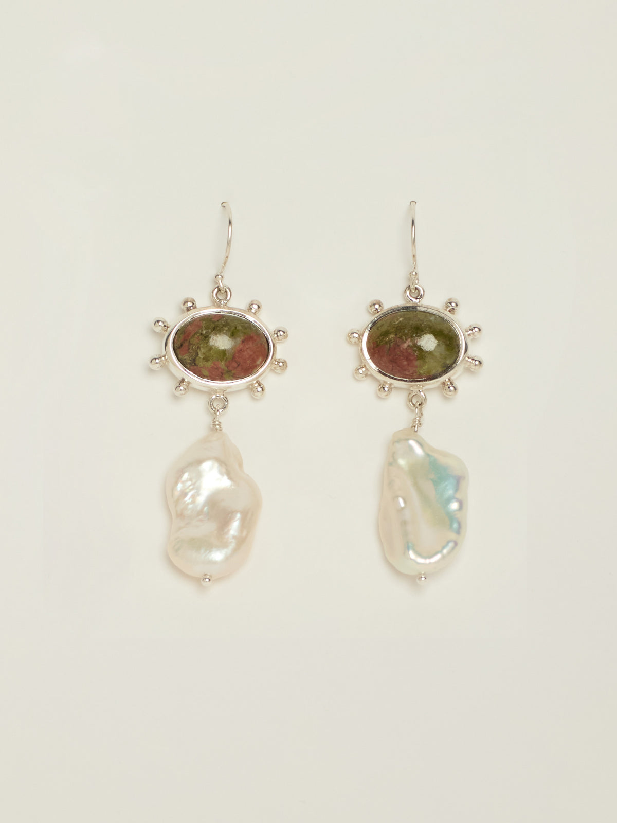 A pair of dangle earrings featuring oval Unakite stones in silver with bobble details around the settings, with irregular white baroque pearls hanging below, shown on a light cream background.