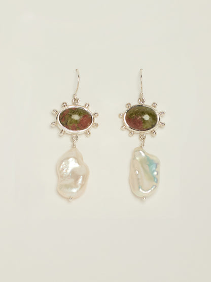 A pair of dangle earrings featuring oval Unakite stones in silver with bobble details around the settings, with irregular white baroque pearls hanging below, shown on a light cream background.