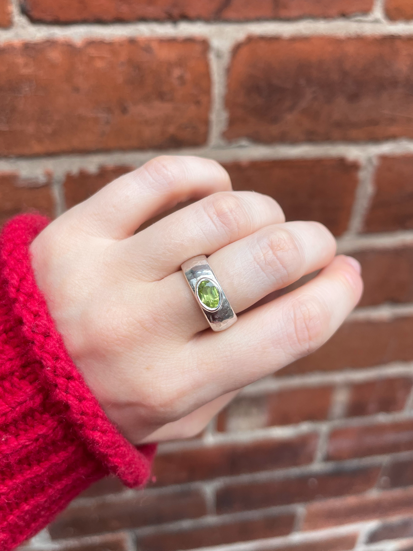 Hand wearing a chunky silver ring with a green peridot against a brick wall