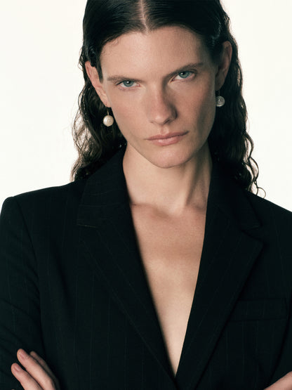 A close-up portrait of a woman with dark hair wearing a black pinstripe blazer and pearl drop earrings against a plain light background.