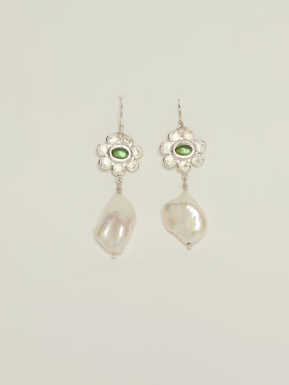 A pair of silver dangle earrings featuring a small oval green tourmaline stone in a floral setting, with a large, irregular white baroque pearl hanging below, shown against a light gray background.