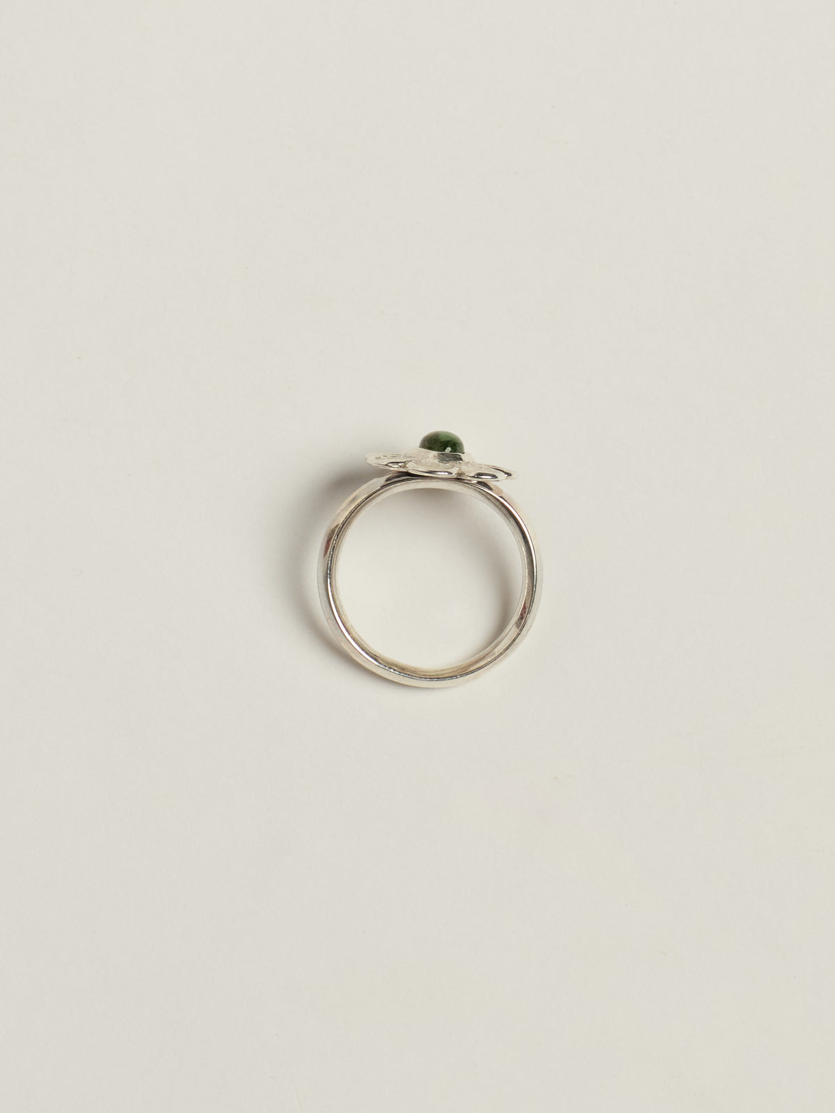 A silver ring with a small green oval gemstone, photographed from above on a light beige background.