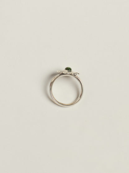 A silver ring with a small green oval gemstone, photographed from above on a light beige background.