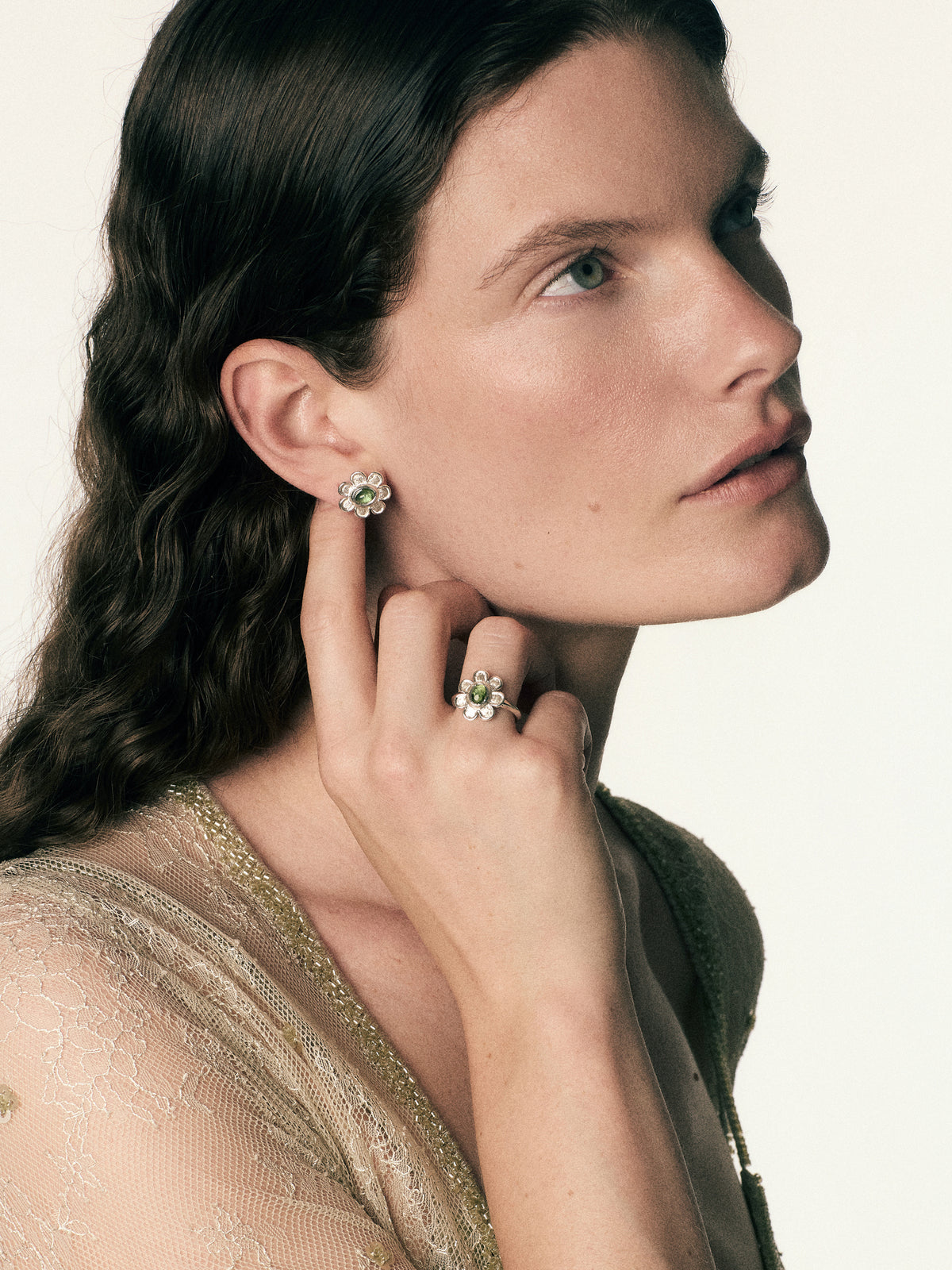A close-up portrait of a woman with dark hair wearing a sparkling floral cluster earring and a matching ring, set against a clean off-white background.