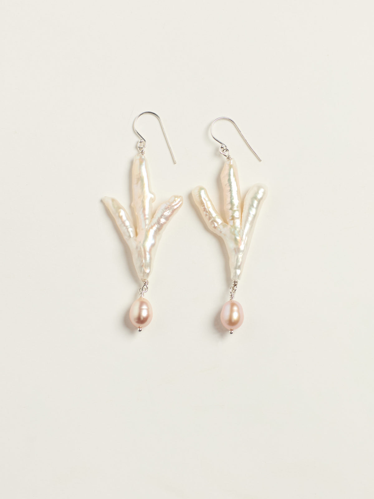A pair of dangle earrings featuring natural baroque pearls arranged in a claw like shape, with a small teardrop pearl hanging below each, suspended from silver fishhook wires on a light beige background.