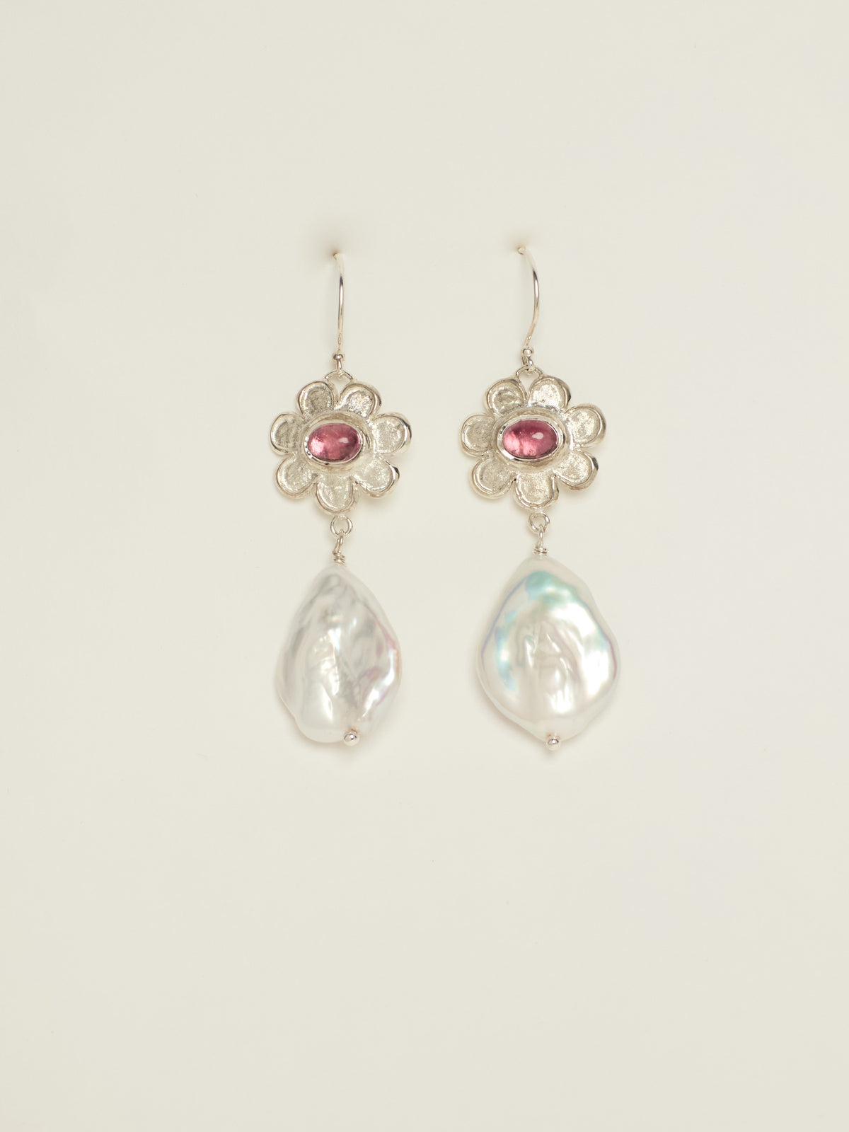 A pair of silver dangle earrings featuring a small silver flower with a pink gemstone and a teardrop-shaped white baroque pearl below, set against a plain off-white background.