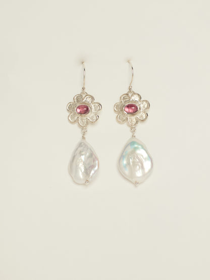 A pair of silver dangle earrings featuring a small silver flower with a pink gemstone and a teardrop-shaped white baroque pearl below, set against a plain off-white background.