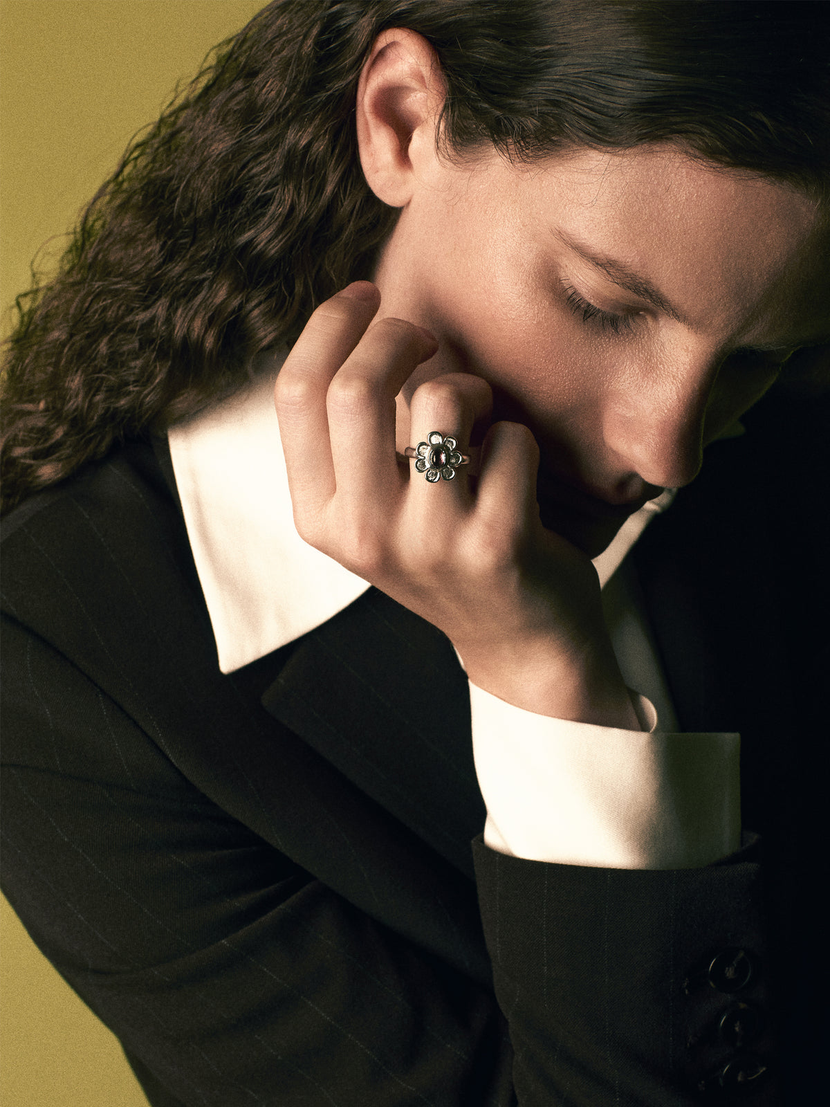 A close-up portrait of a person with long dark curly hair, wearing a black blazer over a white collared shirt, with a silver ring featuring a floral cluster design on their finger.