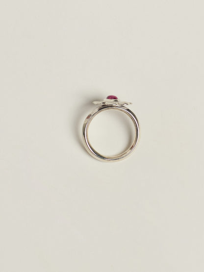 A silver ring featuring a small flower setting with a small pink gemstone, photographed on a clean, light beige background.