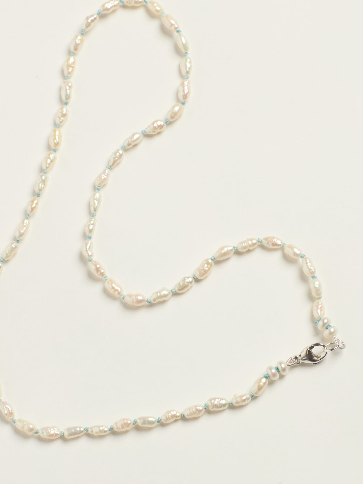 A delicate beaded necklace featuring irregular, pearly off-white baroque pearls separated by thin light blue threads, finished with a silver lobster clasp, laid out on a clean white background.