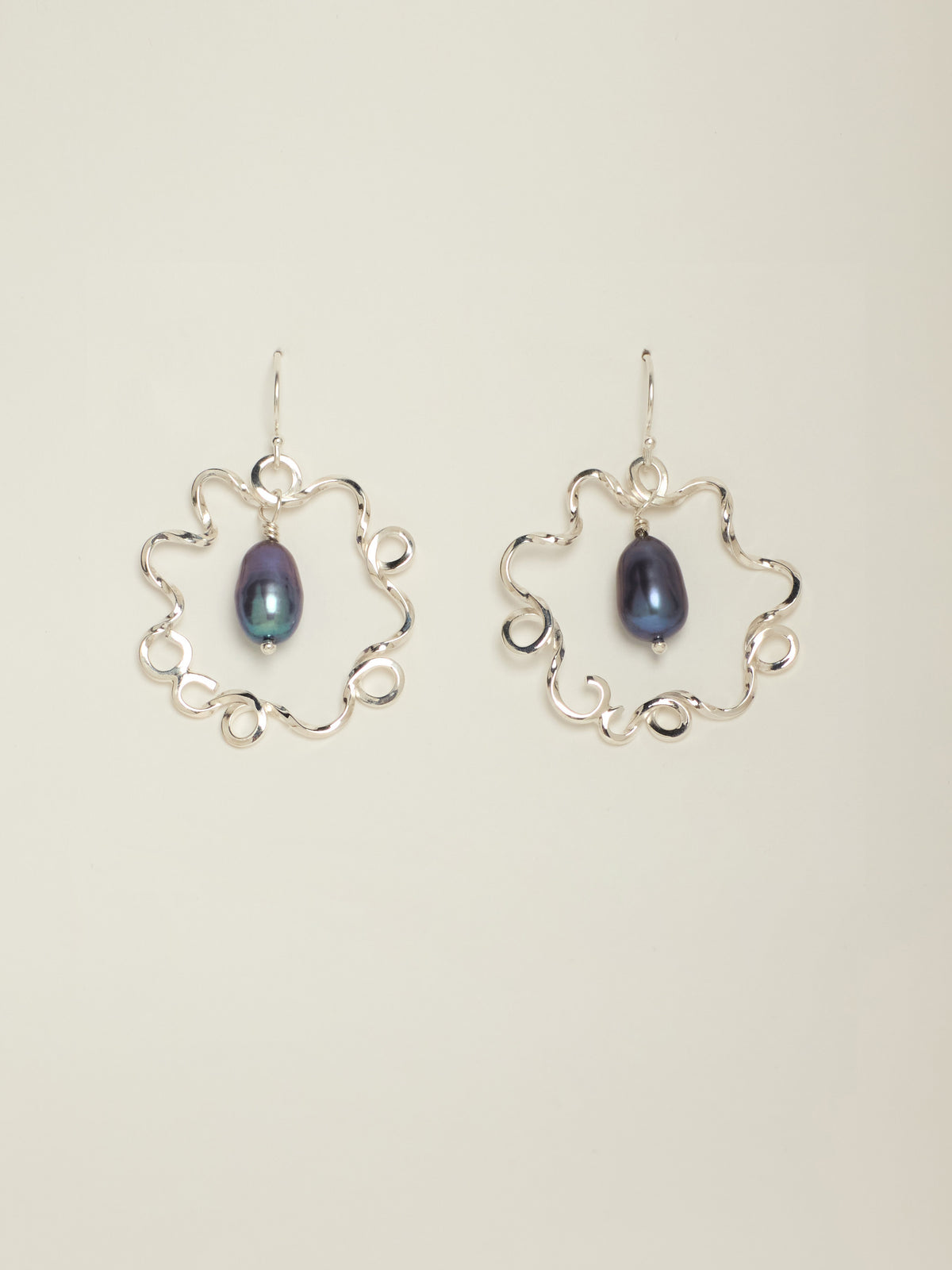 A pair of silver dangle earrings featuring an open, scalloped circular frame with a teardrop-shaped dark blue iridescent pearl in the center, set against a plain light beige background.