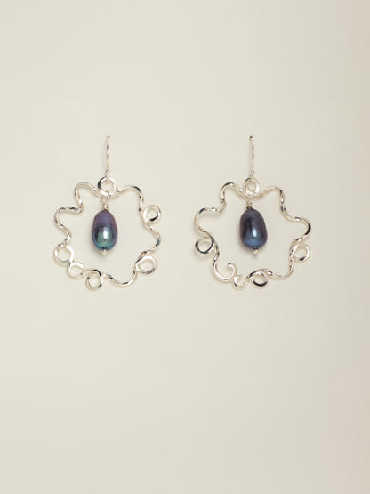A pair of silver dangle earrings featuring an open, scalloped circular frame with a teardrop-shaped dark blue iridescent pearl in the center, set against a plain light beige background.