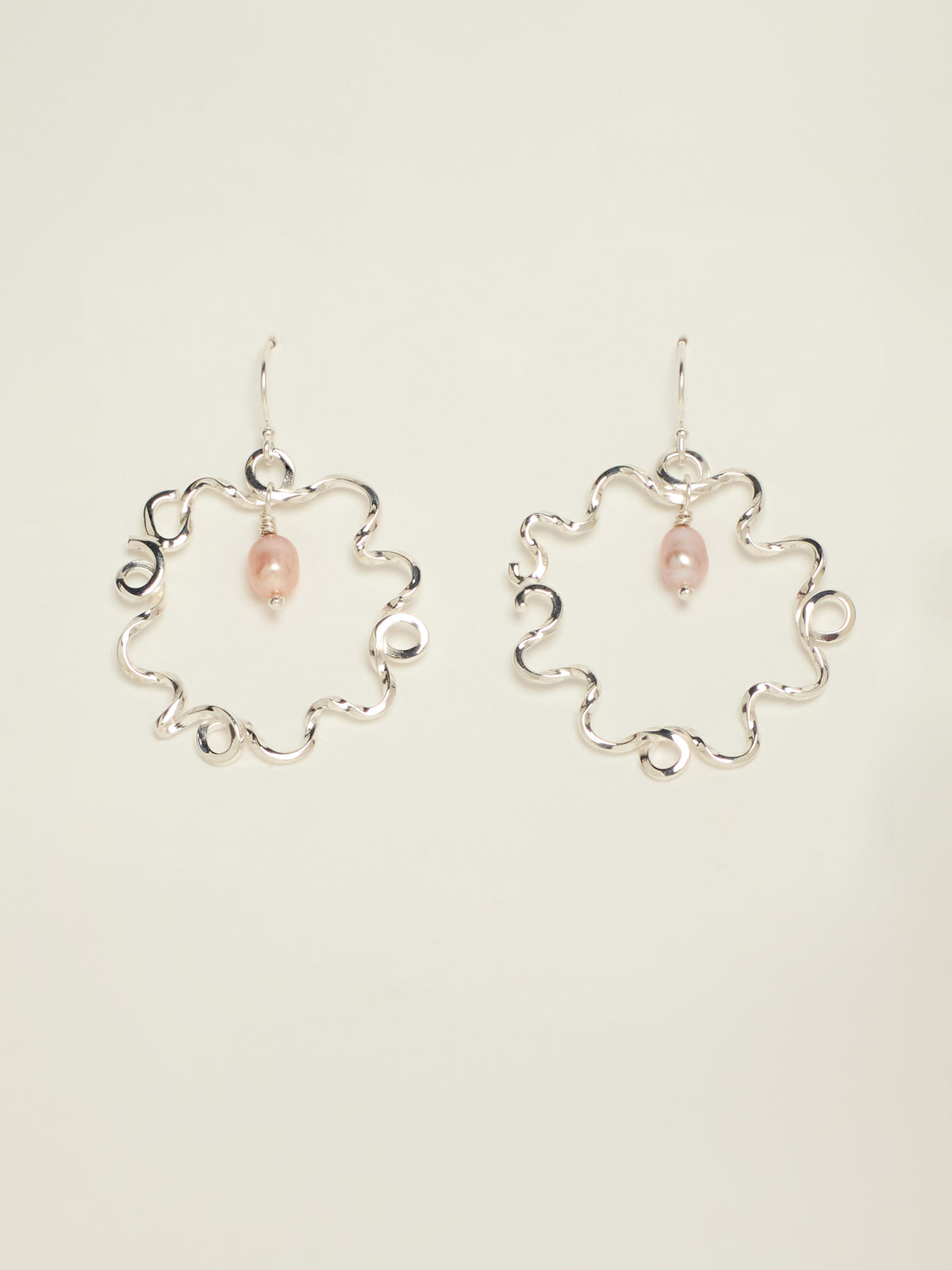 A pair of silver-tone wavy hoop earrings with a small pink teardrop bead hanging from each earring, displayed against a solid off-white background.