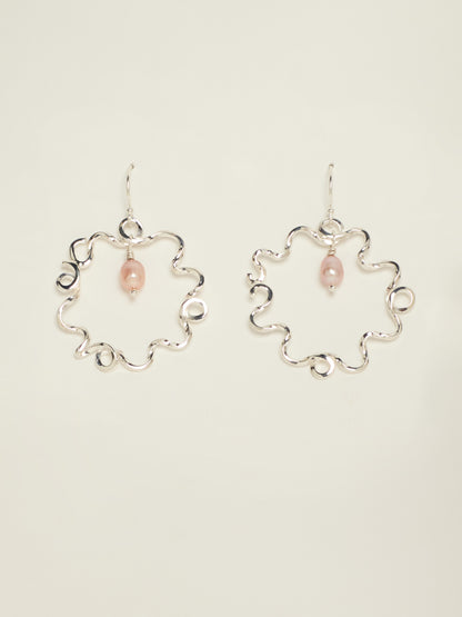 A pair of silver-tone wavy hoop earrings with a small pink teardrop bead hanging from each earring, displayed against a solid off-white background.