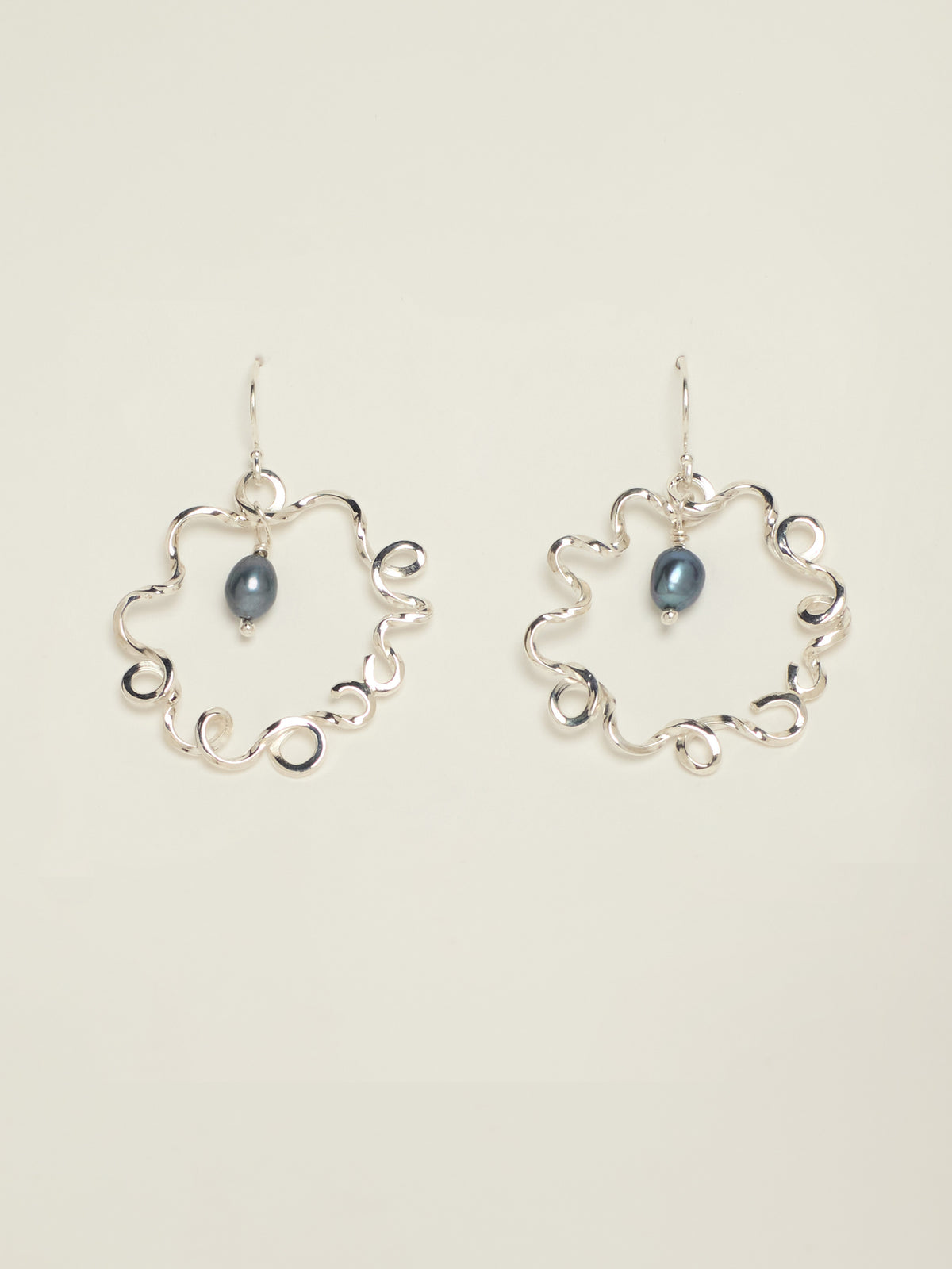 A pair of silver hoop earrings with an open, wavy scalloped circular design and a small dark blue teardrop pearl hanging in the centre of each earring, shown against a solid light beige background.