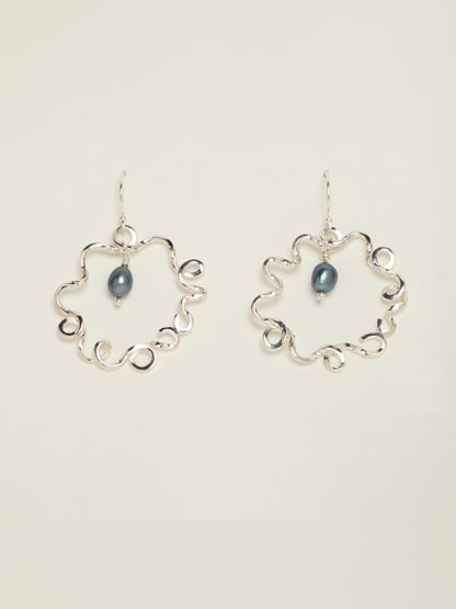 A pair of silver hoop earrings with an open, wavy scalloped circular design and a small dark blue teardrop pearl hanging in the centre of each earring, shown against a solid light beige background.