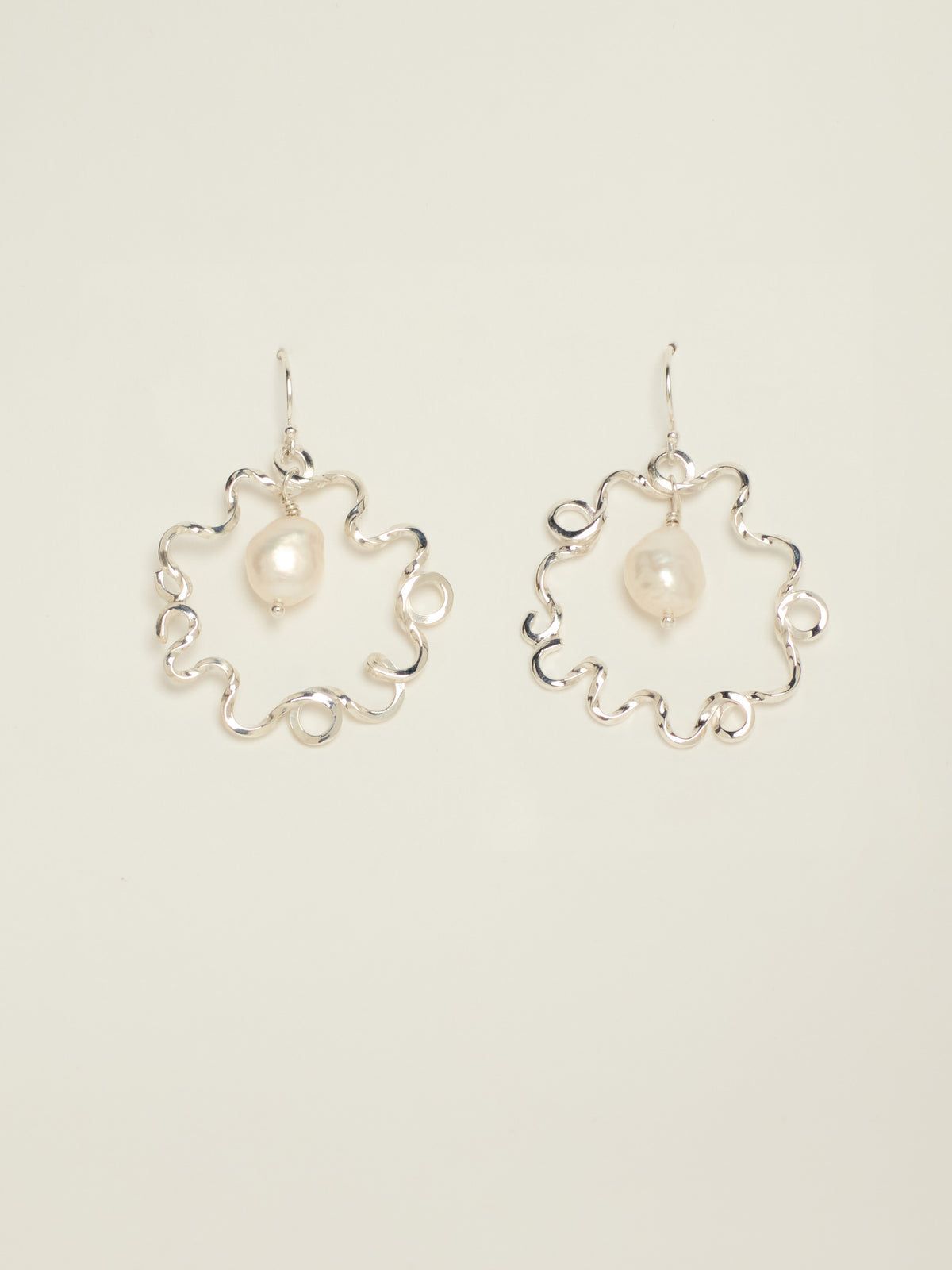 A pair of silver-tone wavy hoop earrings, each featuring a small, irregular white pearl at the center, displayed against a solid off white background.