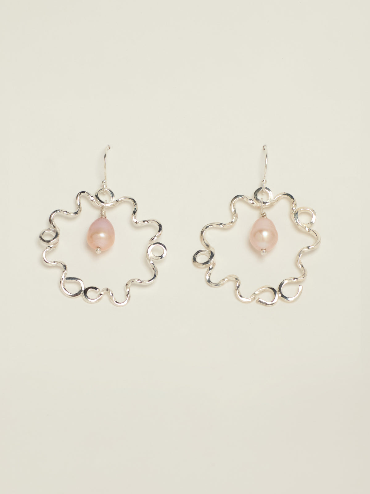 A pair of silver dangle earrings with a wavy openwork circular frame and a small teardrop shaped pink pearl hanging in the centre, displayed on a light beige background.