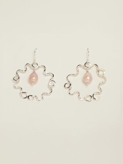 A pair of silver dangle earrings with a wavy openwork circular frame and a small teardrop shaped pink pearl hanging in the centre, displayed on a light beige background.
